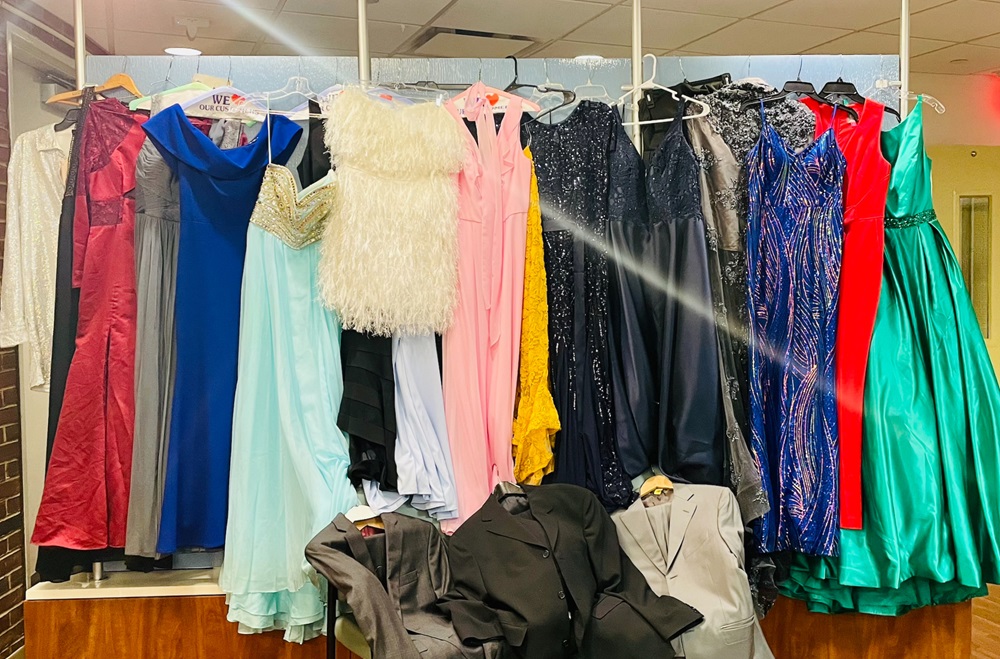 A row of colorful prom dresses hangs in the background, with 3 men’s suits displayed in front 